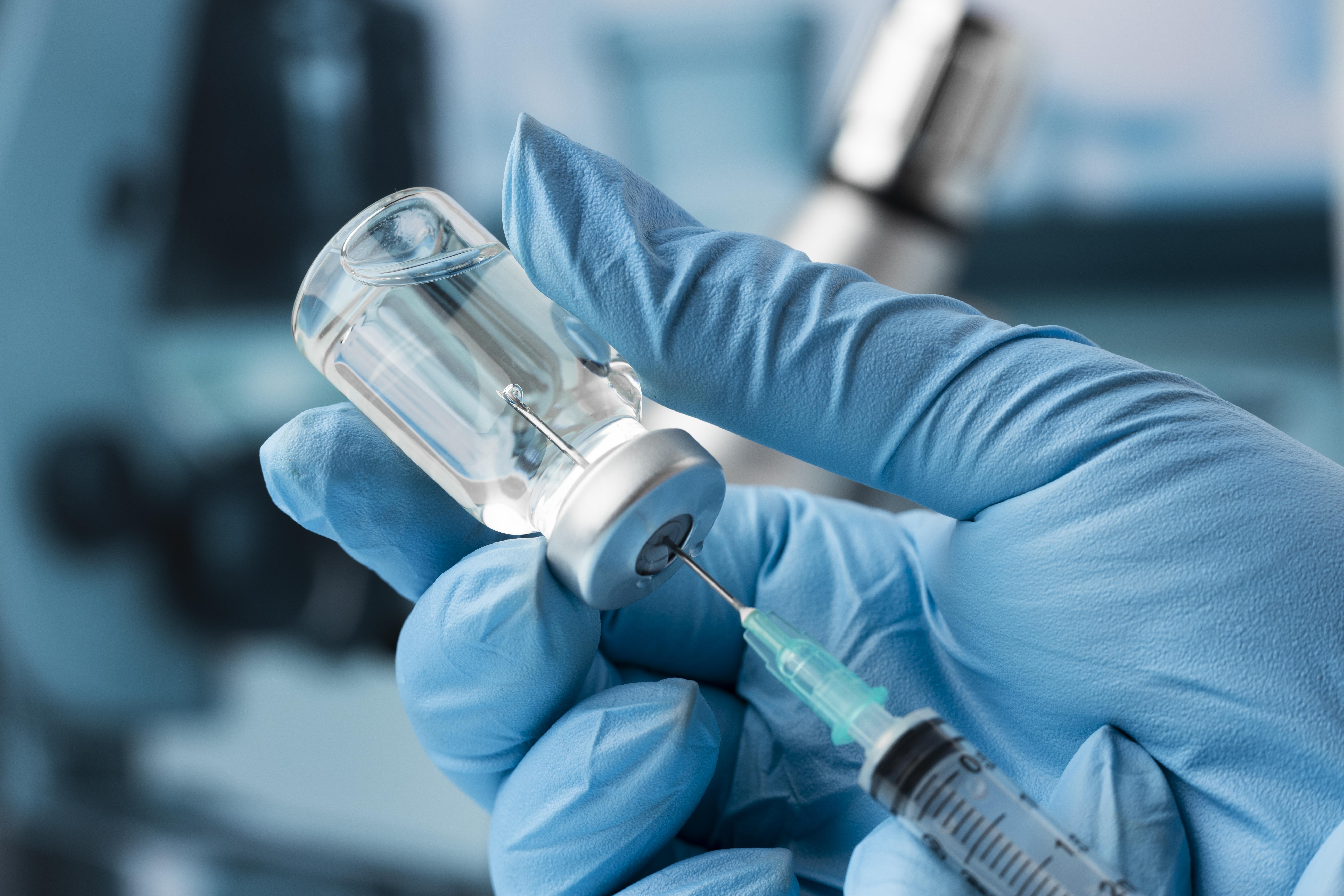 Mão de um profissional de saúde com luva cirúrgica azul usando uma seringa para retirar vacina de uma ampola